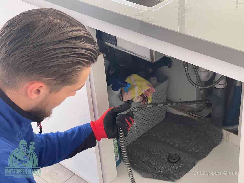 Een monteur voert een flexibele ontstoppingsveer in onder een keukenkastje om een verstopping voorbij de sifon te bereiken in Almelo