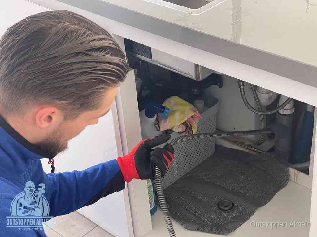 Een monteur voert een flexibele ontstoppingsveer in onder het keukenkastje, langs de sifon, om de gootsteen te ontstoppen in Almelo