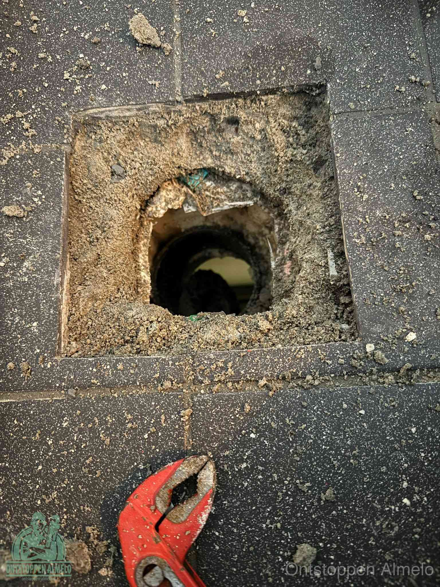 Een geopend rioolputje in een betontegelvloer met losgemaakt rooster en een zichtbare ronde afvoerbuis, het achterhalen waar de afvoerverstopping zit in Almelo