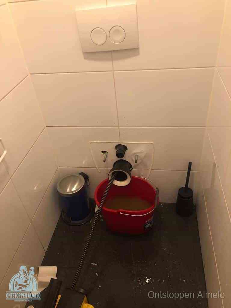 Een ontstopte wand-wc met verwijderde toiletpot waar een ontstoppingsveer in de wanduitgang verdwijnt, het achterhalen waar de verstopping zit in Almelo