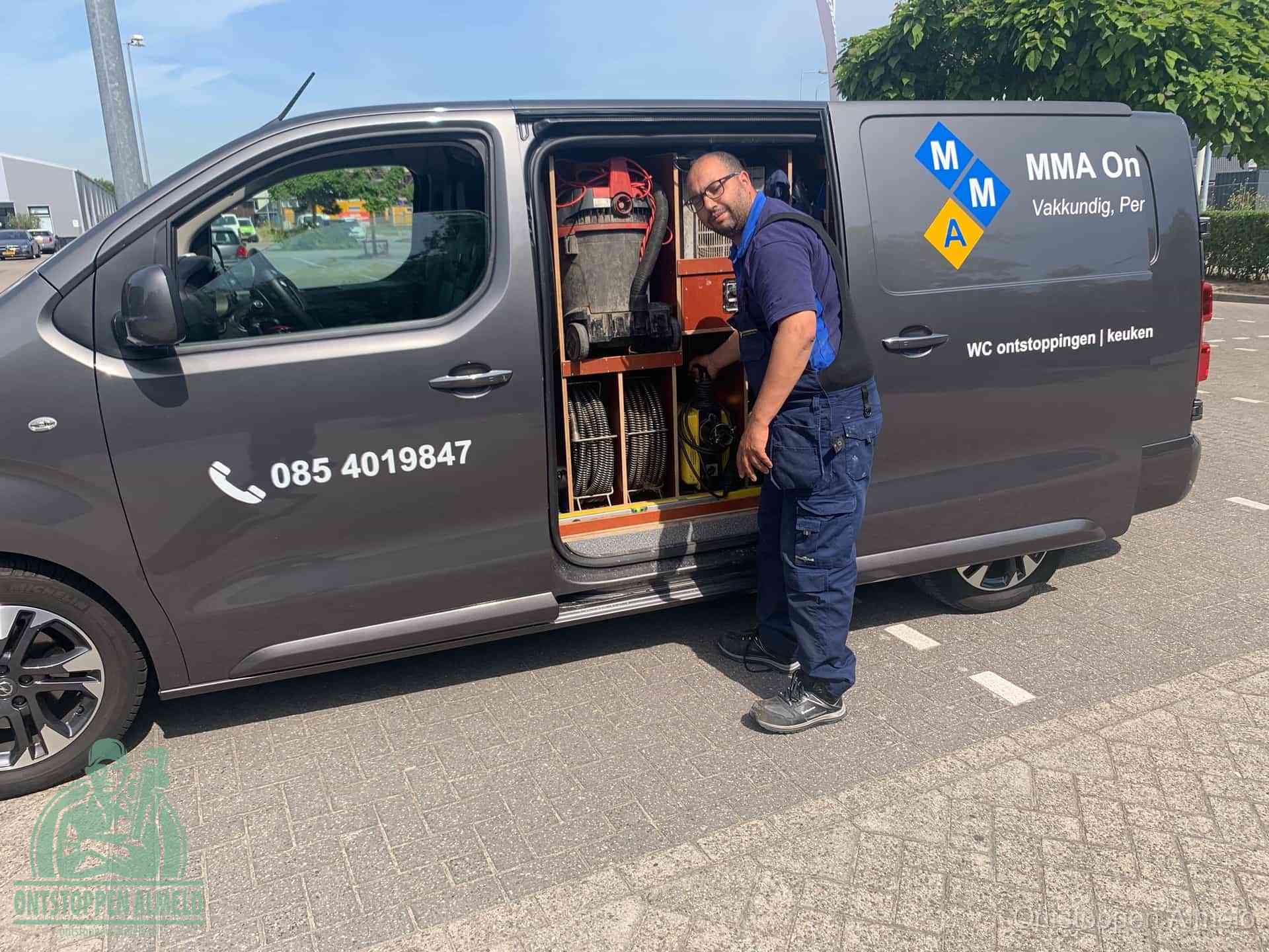 Een monteur bij de open schuifdeur van zijn bedrijfsbus op een parkeerplaats, klaar om de oorzaak van terugkerende afvoerverstoppingen aan te pakken in Almelo