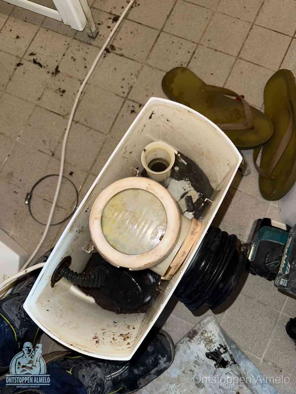 Een gedemonteerde toiletpot op de vloer met ontstoppingsveer en gereedschap ernaast, het verwijderen van een vastzittend voorwerp uit de wc in Almelo