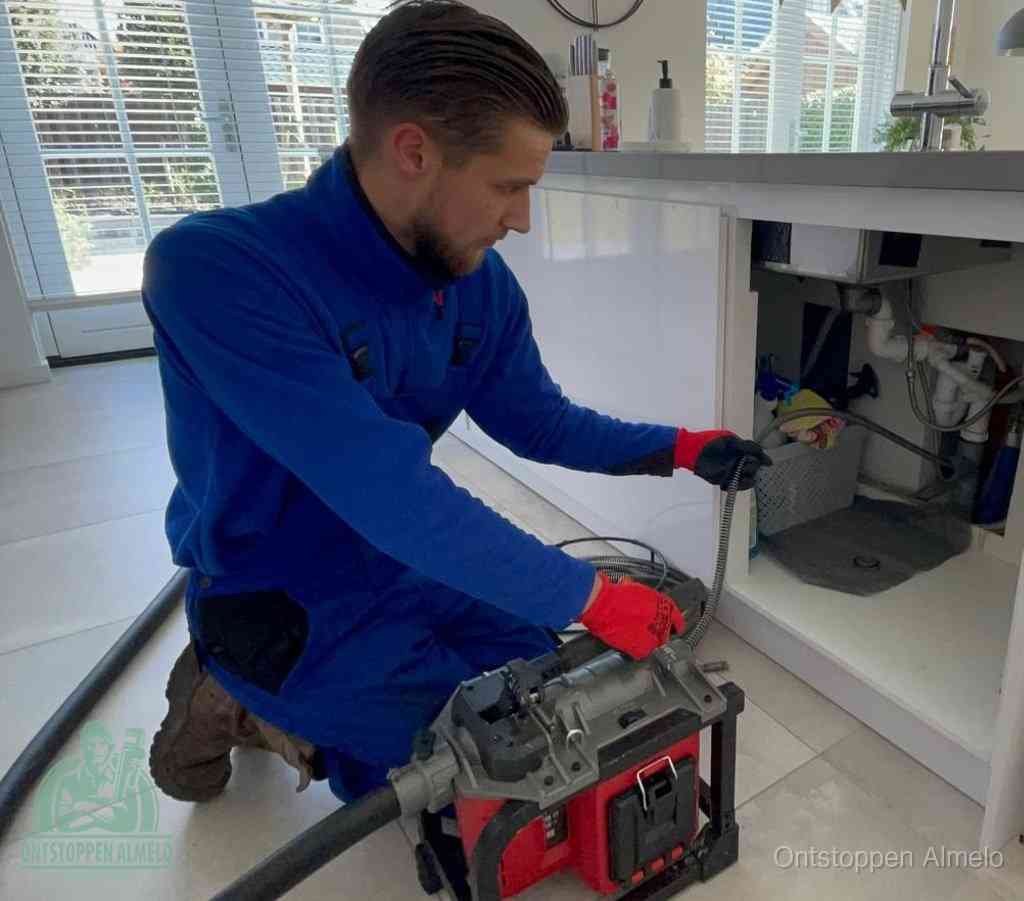 Een monteur met rode handschoenen knielt in een keuken en voert een elektrische ontstoppingsveer in tijdens het professioneel afvoer ontstoppen in Almelo